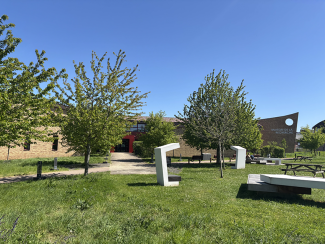 Photo. Au premier plan : un espace vert arboré avec des structures d'assises. Au second plan : le bâtiment de la Maison de la Recherche